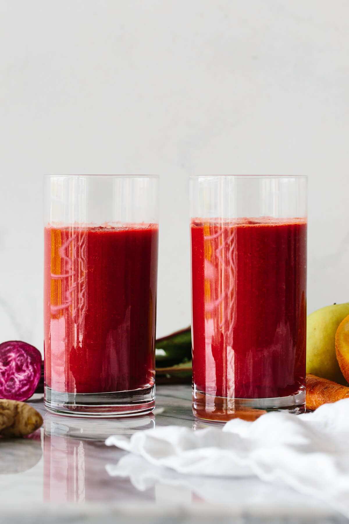 Two glasses of apple, carrot, beet smoothie surrounded by fresh produce.