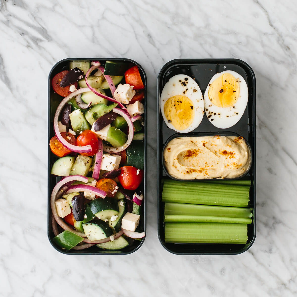 Bento box filled with Greek salad, hard boiled egg, celery and hummus.