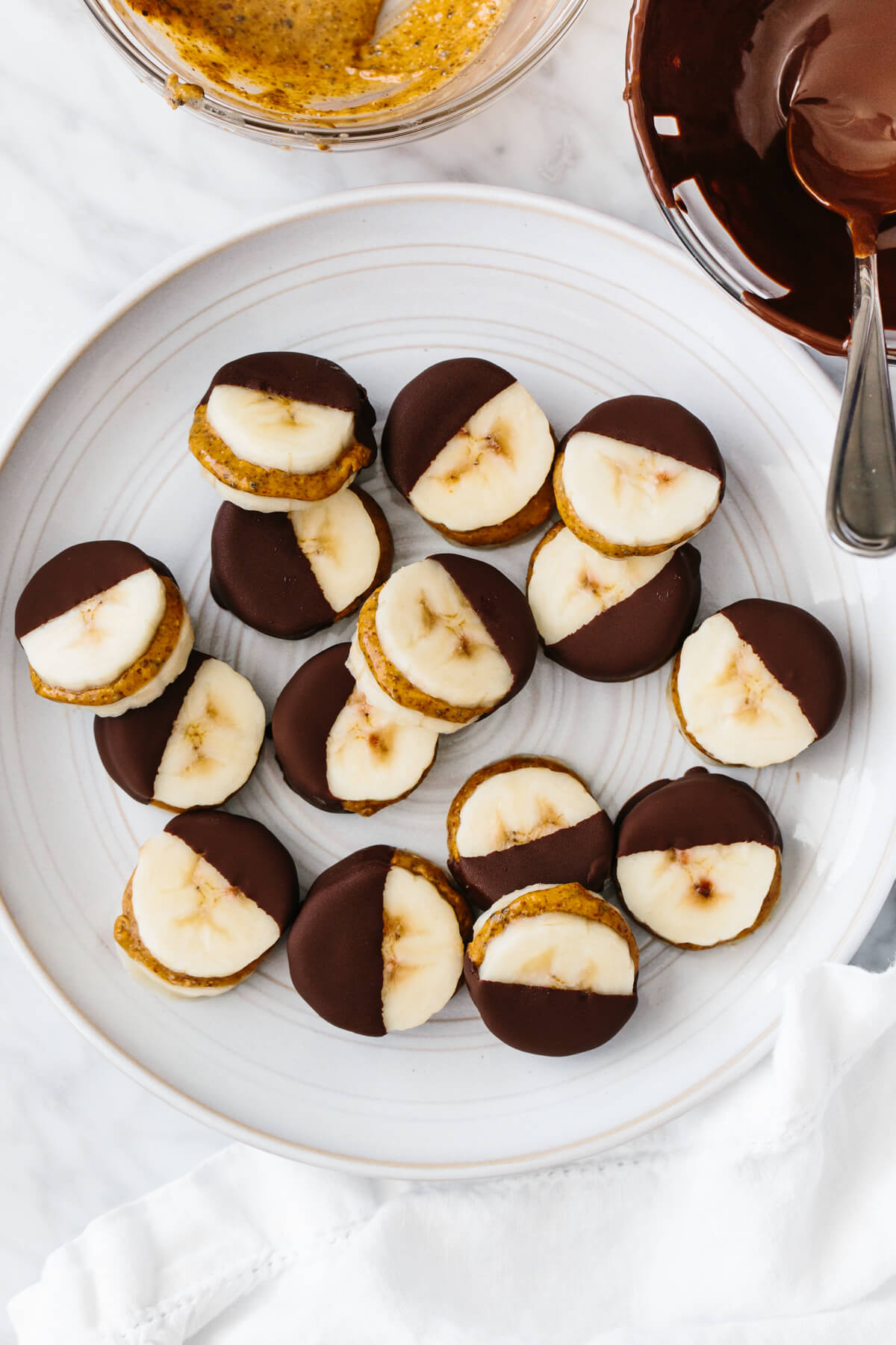 Frozen chocolate banana bites on a plate next to bowls of chocolate and almond butter.
