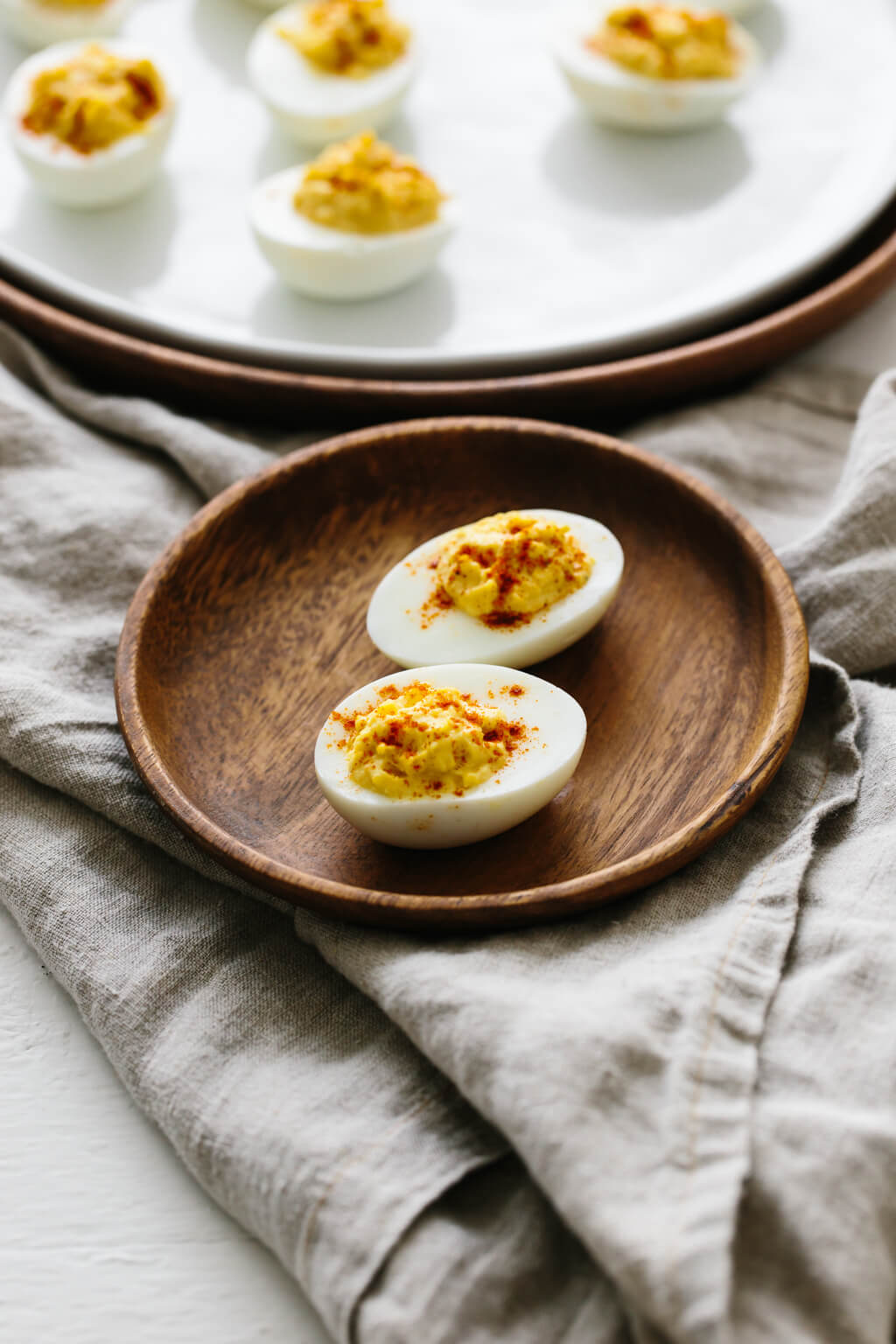 Two deviled eggs on a small wood plate with more deviled eggs in the background. 