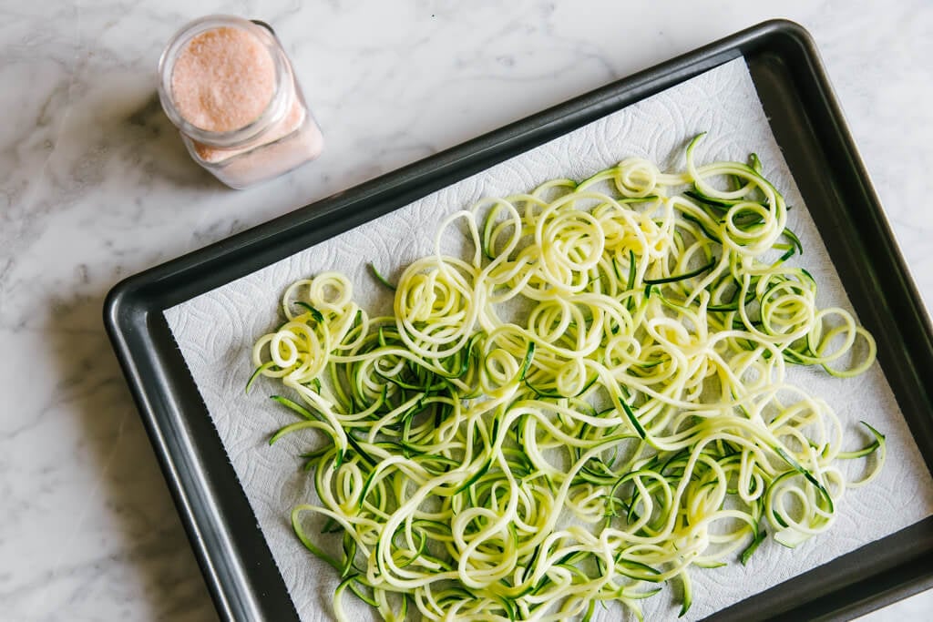 Zucchini noodles on a baking sheet next to a container of salt.