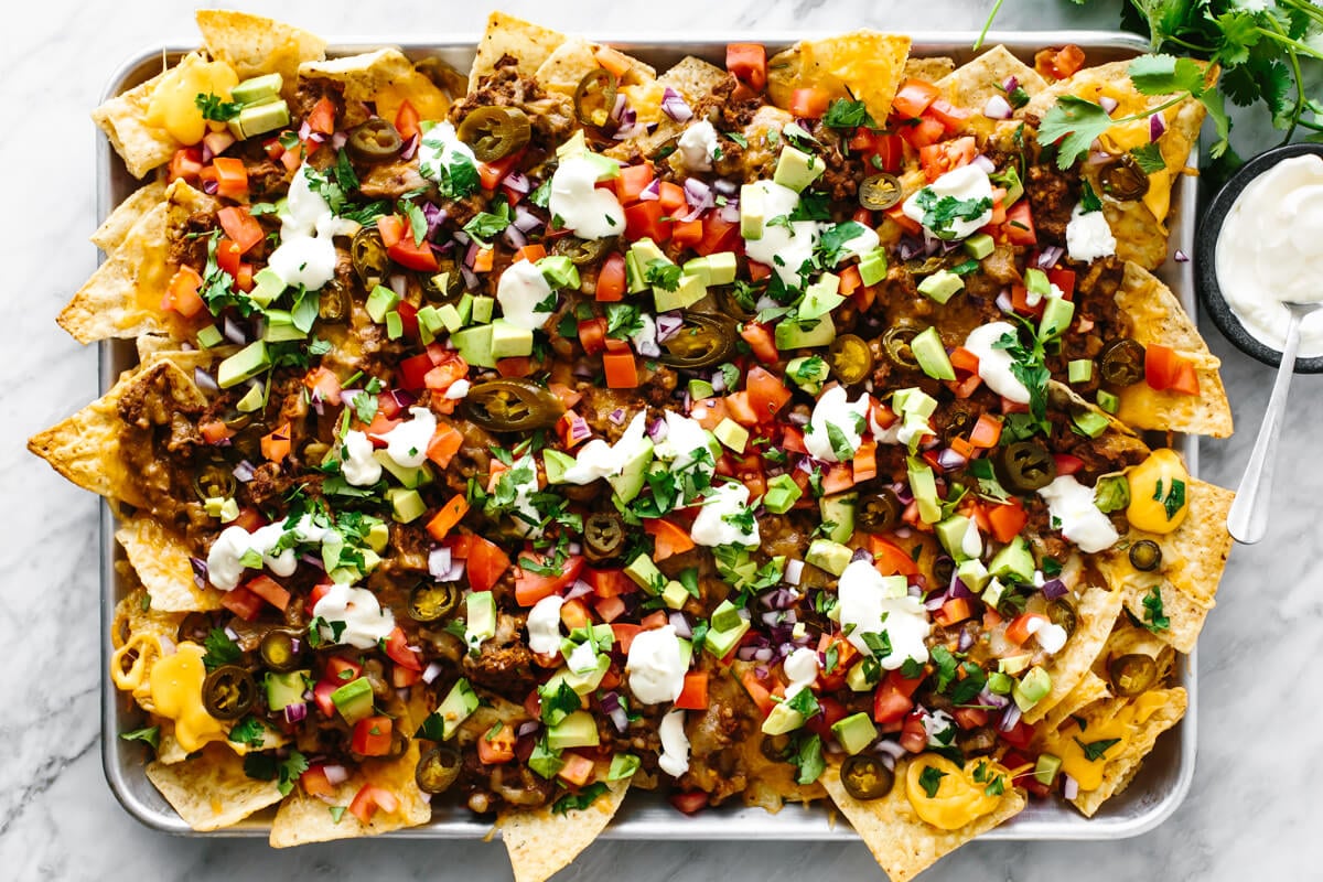 A sheet pan loaded with nachos next to sour cream.