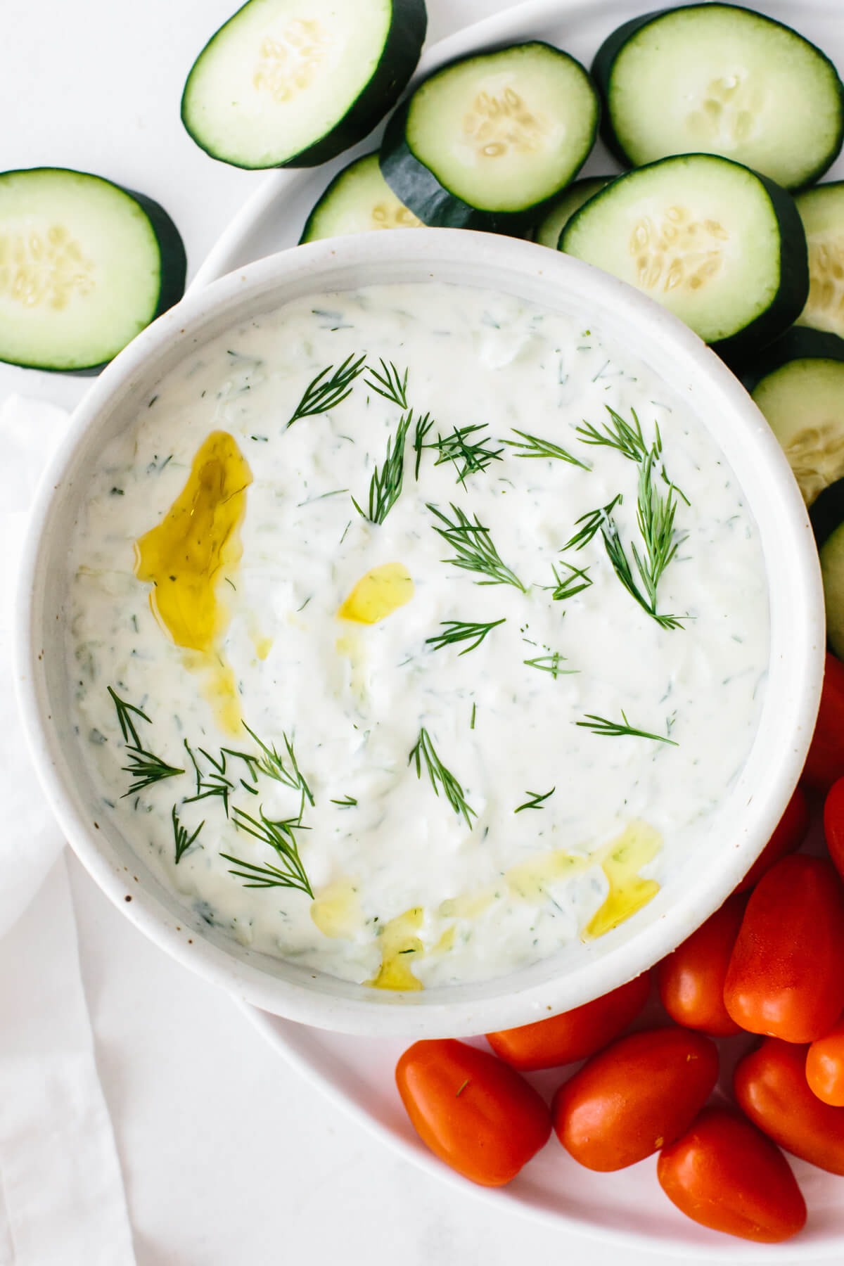 Tzatziki in a bowl surrounded by sliced cucumber and cherry tomatoes.